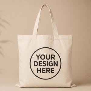 May include: A beige canvas tote bag with two shoulder straps and a centred black circular graphic containing the text YOUR DESIGN HERE in bold capital letters. The bag features a rectangular body with a flat bottom and visible fabric texture.
