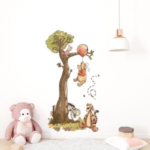 Puede incluir: Una calcomanía de pared caprichosa con Winnie the Pooh, Piglet, Eeyore y Tigger. Pooh cuelga de un globo, mientras que Piglet está sentado en un árbol. Tigger y Eeyore están en la base del árbol. Un oso de peluche rosa está en primer plano.