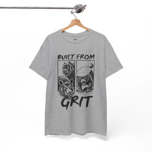 Peut inclure: T-shirt gris avec l'inscription "BUILT FROM GRIT" et des illustrations d'un chien, d'un ours et d'un sanglier. Le t-shirt est suspendu à un cintre en bois.