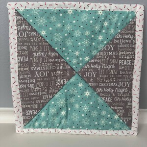 May include: A square quilted fabric coaster with a white background and a red dotted border. The coaster is made of four triangles of fabric, each with a different pattern. The top triangle is a teal blue fabric with white snowflakes and stars. The bottom triangle is a grey fabric with white text that reads "Peace, Let it Snow, Merry & Bright, Oh Holy Night, Believe, Jingle All the Way, Merry Christmas, Joy". The left and right triangles are the same as the top triangle.