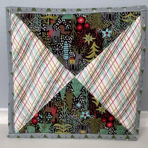 May include: A square quilted hot pad with a grey border and a pattern of plaid and a woodland scene with deer, trees, and flowers.