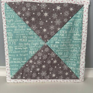 May include: A quilted Christmas-themed wall hanging with a snowflake pattern in grey and white. The fabric is a light blue colour with white text that says "Peace, Joy, Let it Snow, Merry Christmas, Silent Night, Oh Holy Night, and Believe". The border is white with a red dotted line.