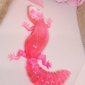 Puede incluir: Una figurita de gecko de resina translúcida rosa con un acabado brillante. El gecko está enroscado con la cola envuelta alrededor de su cuerpo.