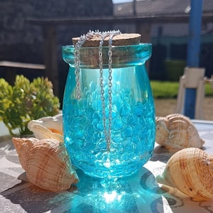 Peut inclure: Un collier en chaîne argentée se trouve dans un bocal en verre bleu rempli de perles bleues. Le bocal a un couvercle en liège et est entouré de coquillages.