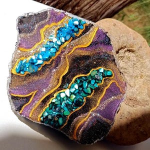 Puede incluir: Una piedra decorativa con diseño de geoda. La piedra presenta una base morada y negra con detalles dorados e incrustaciones de piedras turquesas y verdes. Los bordes son plateados y brillantes, creando una pieza artística única.
