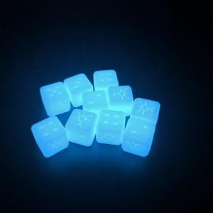 May include: A group of ten glow-in-the-dark dice with white markings. The dice are a light blue colour and are arranged in a pile.