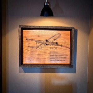 Puede incluir: Arte de pared enmarcado en madera con un diseño de avión grabado. La obra de arte incluye la palabra "PILOT" y una definición. El marco es de color marrón oscuro y la madera tiene una veta natural.