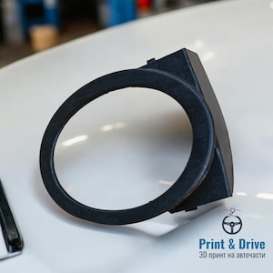 以下が含まれることがあります： 黒色の3Dプリントされた自動車部品。円形のリングと長方形のベースが特徴です。車両のインテリアに適合するように設計されています。画像には「Print & Drive」と「3Dプリントの自動車部品」というテキストが含まれています。