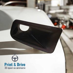 Puede incluir: Una pieza de coche negra impresa en 3D, probablemente una ventilación o una toma de aire, se muestra en un coche blanco. La pieza tiene una superficie lisa y redondeada y un pequeño agujero en el centro. La imagen se toma desde una perspectiva de primer plano, con la pieza en foco y el coche en el fondo.