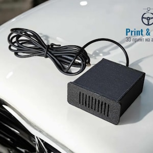 May include: A black, rectangular device with a speaker grille on the front. It is connected to a black cable and is sitting on a white car hood. The text "Print & Drive 3D print na avtochasti" is visible in the upper right corner of the image.