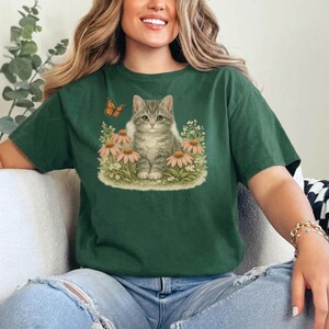 Peut inclure: T-shirt vert foncé avec un chaton tigré gris assis au milieu de fleurs roses et de verdure. Un papillon orange vole près du chaton. Le t-shirt est en matière souple.