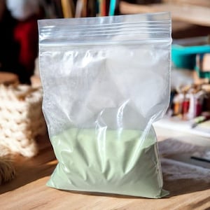 Può includere: Un sacchetto di plastica trasparente richiudibile riempito con polvere verde chiaro. Il sacchetto è su una superficie di legno.