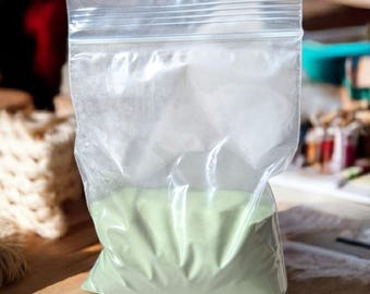 May include: A clear plastic resealable bag filled with a light green powder. The bag is sitting on a wooden surface.