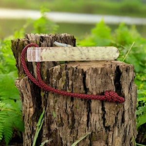 Puede incluir: Una varita de cristal rectangular de color claro con una cuerda roja adjunta, apoyada en un tocón de árbol. La varita tiene un patrón moteado. La cuerda roja está anudada en el extremo. El fondo presenta follaje verde y una masa de agua.