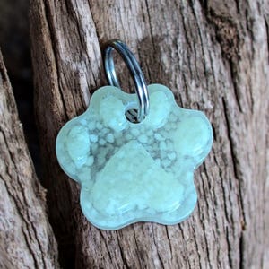 Op de afbeelding: Een lichtgroen, pootvormig huisdierlabel met een zilveren metalen ring. Het label heeft een gespikkeld ontwerp en is geplaatst tegen een verweerde houten achtergrond. Het label is ontworpen voor huisdieren.