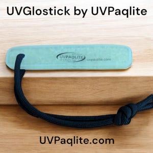 Peut inclure: Un UVGloStick bleu clair par UVPaqlite avec un cordon noir. Le UVGloStick est un petit objet rectangulaire avec une surface texturée. Le cordon est noué. Le texte "UVPaqlite.com" est imprimé sur le UVGloStick et sur la surface en bois en dessous.