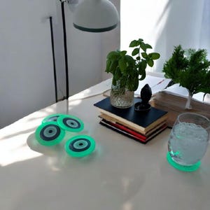 Könnte beinhalten: Vier grüne Glasuntersetzer mit einem blauen bösen Auge Design. Die Untersetzer befinden sich auf einem weißen Tisch mit einem Glas Wasser und einem Stapel Bücher.