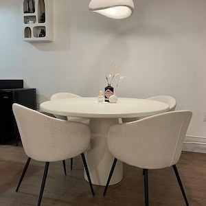 Könnte beinhalten: Ein modernes Esszimmer-Set mit einem runden weißen Tisch und vier cremefarbenen Stühlen mit schwarzen Beinen. Auf dem Tisch stehen eine Vase mit Trockenblumen und eine Flasche. Überkopf hängt eine einzigartige Leuchte von der Decke. Der Tisch hat etwa 120 cm Durchmesser.