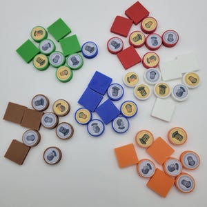 May include: A collection of colorful game pieces, including square tiles and circular tokens. The tiles are in green, red, blue, white, brown, and orange. The tokens feature various illustrations and are arranged in groups, suggesting a board game.