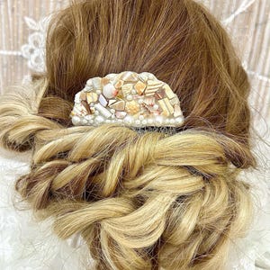 May include: A decorative hair comb featuring a mosaic of seashells in various shapes, sizes, and colors, including white, beige, and brown. The comb is set against a backdrop of styled blonde and brown hair, suggesting a beach or ocean theme.