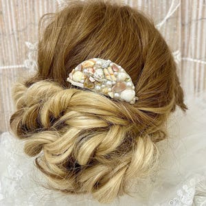 Op de afbeelding: Een decoratieve haar kam met een halve maan ontwerp, versierd met diverse schelpen, parels en kleine versieringen. De kam is geplaatst tegen een achtergrond van gestyled blond en bruin haar, waardoor een accessoire met strandthema ontstaat.
