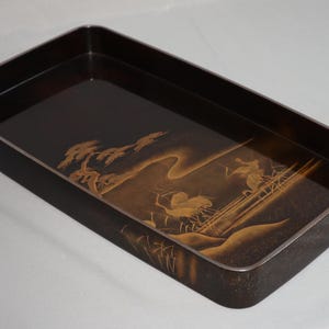 以下が含まれることがあります： 詳細な金色の風景デザインが施された、長方形の濃い茶色のトレイ。トレイは角が丸く、銀色のトリムが付いています。アートワークは、木、人物、橋を描いています。