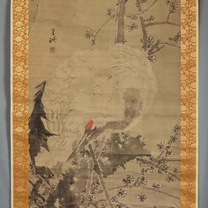 May include: A vertical hanging scroll painting on a tan background featuring a white crane with a red crown perched on a dark tree branch. The crane is depicted in profile with black wing feathers. Plum blossoms surround the bird, and black ink calligraphy with a red seal is visible on the left side.