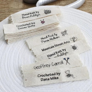 May include: A stack of off-white fabric labels with black text and small illustrations. The labels read "Hand Knit by Bruce Pulitzer", "Crocheted by Grover Dolly", "Hand Knit by Prima Hudson", "Mountain Street Arts", "Geoffrey Carroll", and "Crocheted by Dana Mike".
