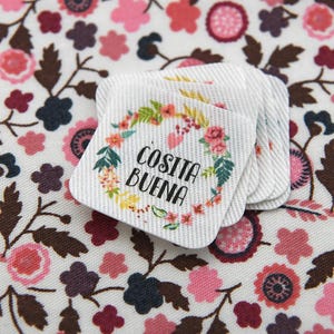 Könnte beinhalten: Ein Stapel quadratischer weißer Stoffetiketten mit abgerundeten Ecken, die ein Blumenkranzdesign in Rosa, Grün und Gelb um den schwarzen Text 'COSITA BUENA' zeigen. Die Etiketten liegen auf einem weißen Stoffhintergrund mit Blumenmuster.