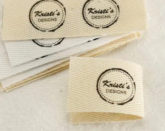 Pode incluir: Uma pilha de etiquetas retangulares de tecido na cor off-white com padrão de espinha de peixe. Cada etiqueta exibe um carimbo circular de tinta preta com o texto Kristi's DESIGNS centralizado. As etiquetas estão dispostas em uma pilha.