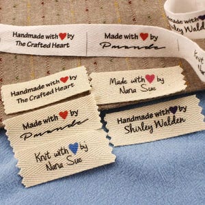 Op de afbeelding: Crèmekleurige stoffen labels met zwarte tekst en harticonen. De labels luiden "Handmade with by The Crafted Heart", "Made with by Pmande", "Made with by Nana Sue", "Handmade with by Shirley Walden" en "Knit with by Nana Sue".
