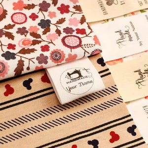 Könnte beinhalten: Nahaufnahme von Stoffen mit Blumen- und geometrischen Mustern. Ein weißes Etikett mit einer Nähmaschinenillustration und dem Text "HANDMADE Your Name" ist angebracht. Weitere Etiketten mit dem Text "Hand made by Your Name" sind ebenfalls sichtbar.