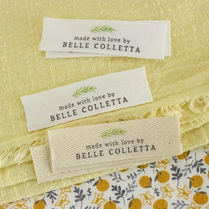 Puede incluir: Tres etiquetas rectangulares de tela color blanco hueso con textura de espiga. Cada etiqueta presenta un pequeño gráfico de hoja verde sobre el texto impreso en negro made with love by BELLE COLLETTA en fuente serif.