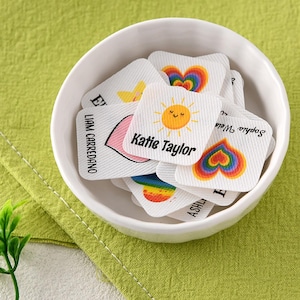 May include: A white ceramic bowl filled with small, square fabric labels. Each label features a unique name and a colourful graphic, such as a smiling yellow sun or a rainbow heart. The text includes names like Katie Taylor and Liam Carredano.