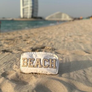 Puede incluir: Bolsa blanca con cremallera con la palabra "BEACH" en letras doradas de purpurina en una playa de arena.