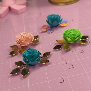 May include: Four paper flower embellishments in shades of teal, peach, and green, each with gold-toned leaves. The flowers are arranged on a pink grid surface. A candle with the text "TOASTED VANILLA" is in the background.