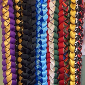 Pode incluir: Uma coleção de colares de fita trançada coloridos, pendurados em um rack. Os colares são feitos com várias cores e padrões, incluindo roxo, dourado, preto, azul, branco, verde, vermelho e amarelo.