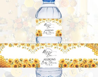 Op de afbeelding: Een heldere waterfles met een blauwe dop en een etiket met zonnebloemen en de tekst "Mommy to Bee Allison's Baby Shower 07.06.23". Het etiket is omgeven door een bloemenrand.