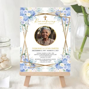 May include: A memorial card on a small wooden easel. The card features a photo of an elderly woman, surrounded by a gold geometric frame and floral accents in blue and white. Text includes the name, dates, and service details.
