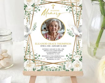 May include: A memorial card on a wooden easel. The card features a photo of a woman, surrounded by white roses, greenery, and a gold geometric frame. Text includes "In Loving Memory" and the woman's name, birth, and death dates.