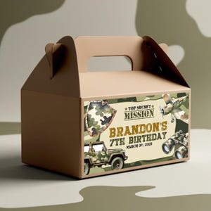 Könnte beinhalten: Eine braune Papp-Geschenkbox mit Griff. Die Box hat ein Tarnmuster mit militärischen Illustrationen, darunter ein Jeep, ein Flugzeug und ein Fernglas. Auf der Box steht "Top Secret Mission" und "Brandons 7. Geburtstag, 09. März 2025."
