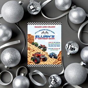May include: A birthday party invitation with a monster truck theme. The invitation features colorful monster trucks racing on a dirt track, with the text "SMASH AND CRASH!" and "ELIJAH'S 5TH BIRTHDAY BASH!" Silver baubles and ribbon surround the invitation.