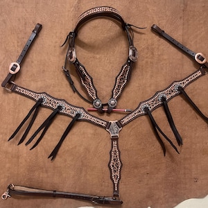 May include: A brown leather horse tack set with leopard print accents, copper-colored hardware, and decorative silver-toned studs. The set includes a headstall, breast collar, and reins, all arranged on a brown surface.