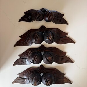 May include: Four dark brown wooden wall carvings, each featuring a pair of spherical fruit or nuts nestled between two carved leaves. The wood displays a smooth, polished finish with visible grain patterns and natural variations in the dark brown tone.