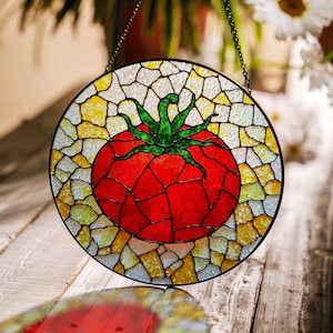 May include: A stained glass window hanging from a chain, depicting a red tomato with a green stem and leaves. The tomato is surrounded by a mosaic of yellow, white, and clear glass.