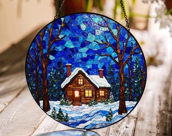 Op de afbeelding: Een hangende ornament van gekleurd glas dat een winterscène voorstelt met een klein huisje in een besneeuwd bos. Het huisje heeft een verlicht raam en een besneeuwd dak. De achtergrond is een blauwe en witte mozaïek.
