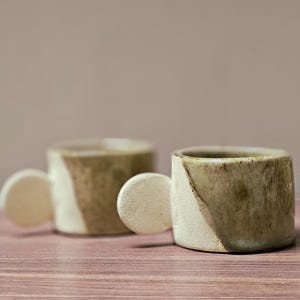 Peut inclure: Deux tasses en céramique au design unique. Chaque tasse a un corps cylindrique avec une poignée arrondie. Les tasses présentent un motif de blocs de couleur diagonaux dans les tons beige et marron. Les tasses sont posées sur une surface en bois.