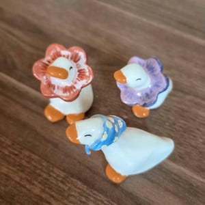 Peut inclure: Trois figurines de canard en céramique. Deux oies ont des couvre-chefs en forme de fleur orange et violet. La troisième oie porte un foulard bleu. Toutes ont des becs et des pieds orange et des corps blancs. Les figurines sont sur une surface en bois.