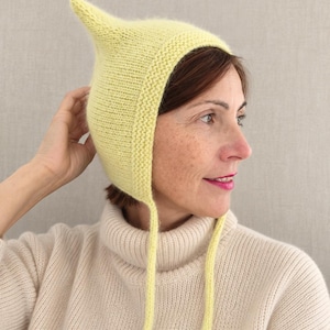 Könnte beinhalten: Eine hellgelbe Strickhaube mit spitz zulaufender Oberseite, einem strukturierten kraus rechts gestrickten Rand entlang der Gesichtsöffnung und zwei langen, dünnen Strickbändern, die vom Kinn herabhängen. Die Haube besteht aus weichem, flauschigem Garn.