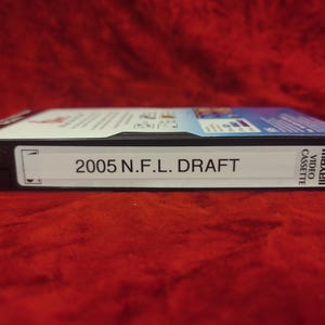 May include: A Maxell video cassette labeled "2005 N.F.L. DRAFT." The cassette is black with a white label and text. The background is a solid red color.